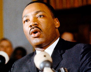Headshot of Rev. Dr. Martin Luther King, Jr., microphone in foreground, out of focus people standing behind him at a distance.